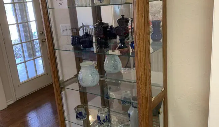 Glass display cabinet with various colored glass vases and pots, situated in a room with a door and hardwood flooring.