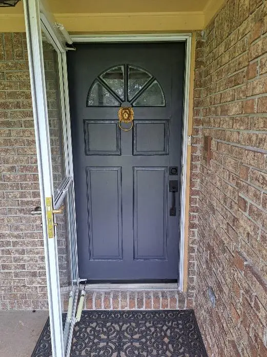 A dark gray front door with a bronze knocker, framed by red brick walls and a white storm door slightly ajar.