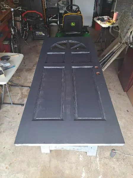 A black painted wooden door with six panels, placed horizontally on a workbench in a cluttered garage.