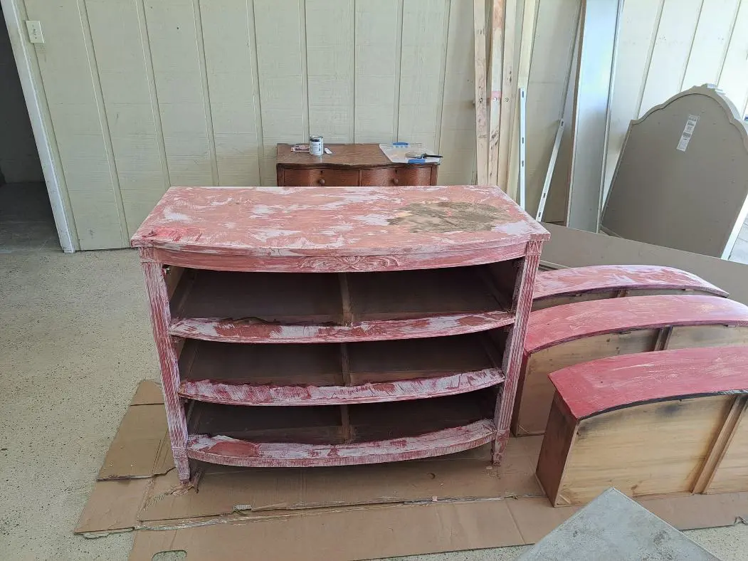 Weathered red and tan wooden cabinet with four shelves, partially peeling paint, in a room with other furniture under renovation.