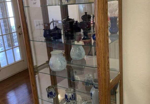 Glass display cabinet with various colored glass vases and pots, situated in a room with a door and hardwood flooring.