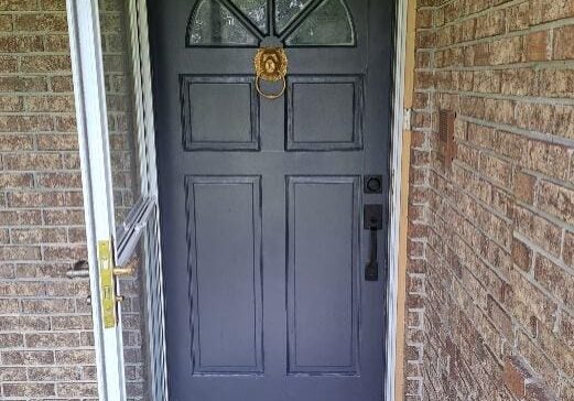 A dark gray front door with a bronze knocker, framed by red brick walls and a white storm door slightly ajar.