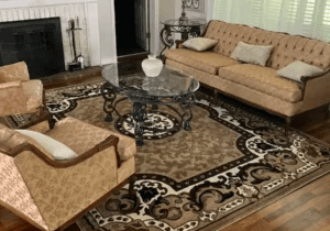 An elegant living room featuring a beige sofa, matching armchair, glass coffee table, ornate rug, and a fireplace with a white mantel.